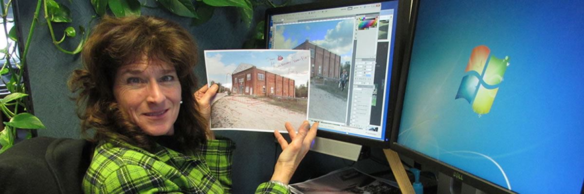 An artist sits in front of her computer screens and holds a printed paper with the source of drawing on the screen